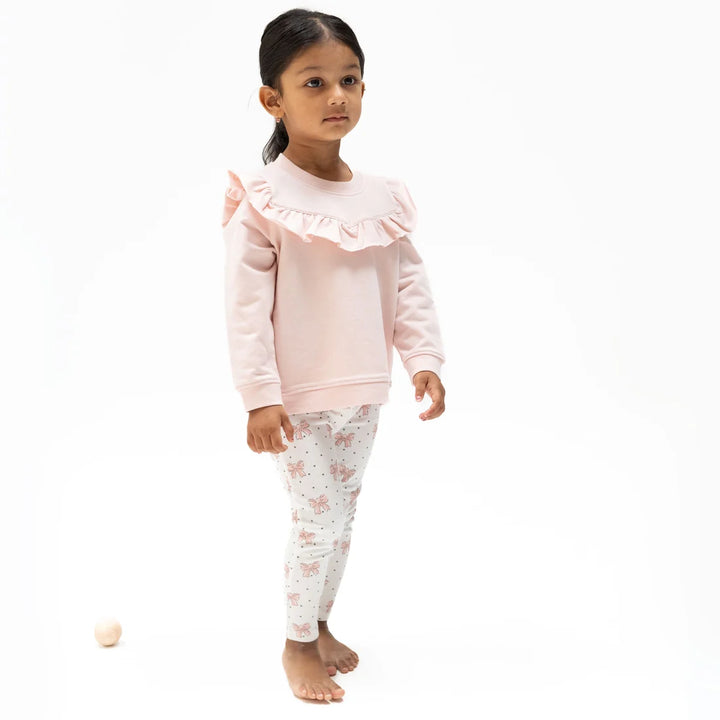 A young girl stands barefoot against a white studio background wearing a light pink long-sleeve sweatshirt with a ruffled collar and white leggings with a pink floral print.