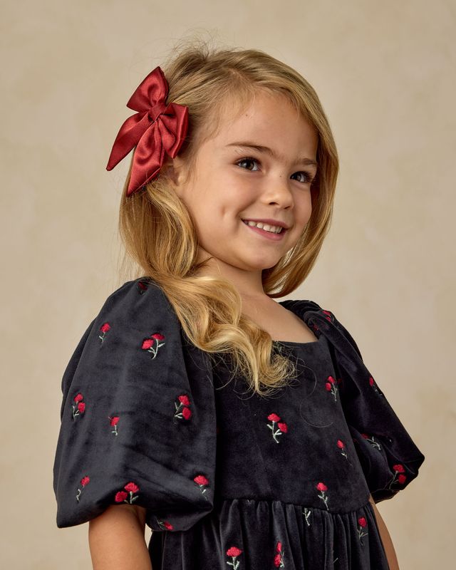 Young girl wearing a black dress with red floral patterns and a large red bow in her hair against a beige background.