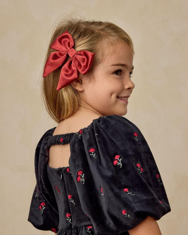 Young girl with a red bow in her hair wearing a black dress with red floral embroidery on a beige background