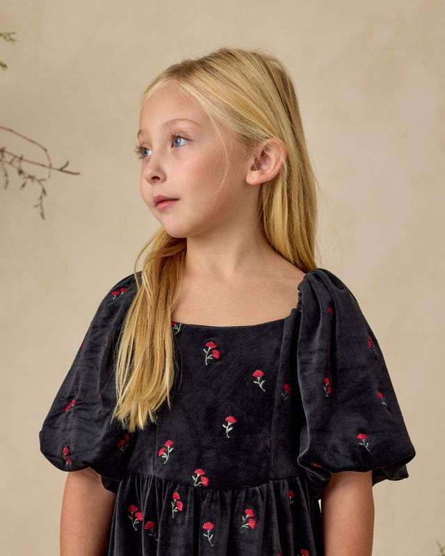 Young girl wearing a black dress with red floral embroidery against a beige background