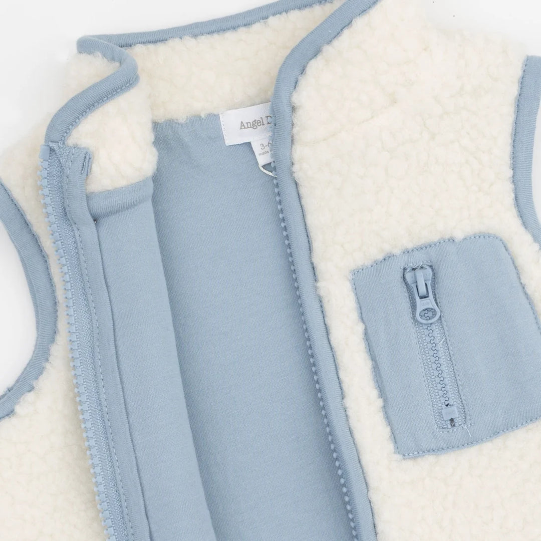 A close-up of a children's zip-front vest with a soft cream sherpa lining and light blue fabric exterior. A front pocket with a matching zipper is visible, along with a neck label that appears to read Angel.