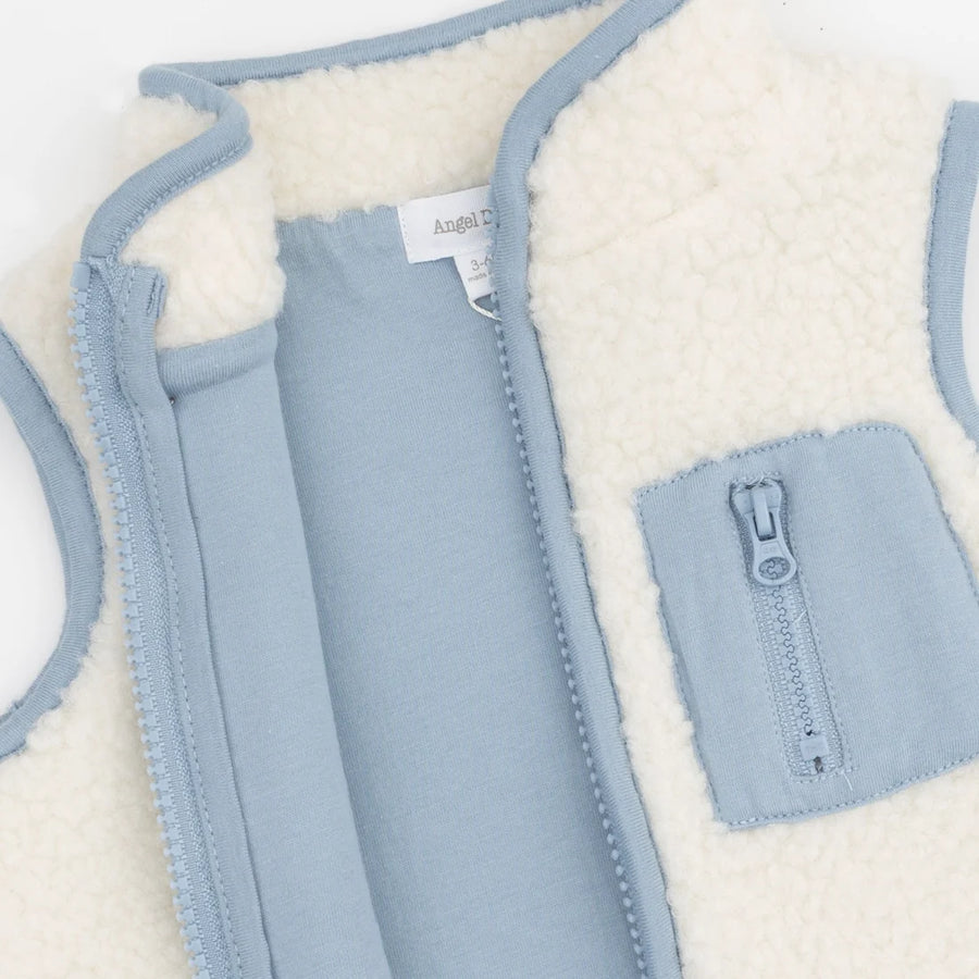 A close-up of a children's zip-front vest with a soft cream sherpa lining and light blue fabric exterior. A front pocket with a matching zipper is visible, along with a neck label that appears to read Angel.