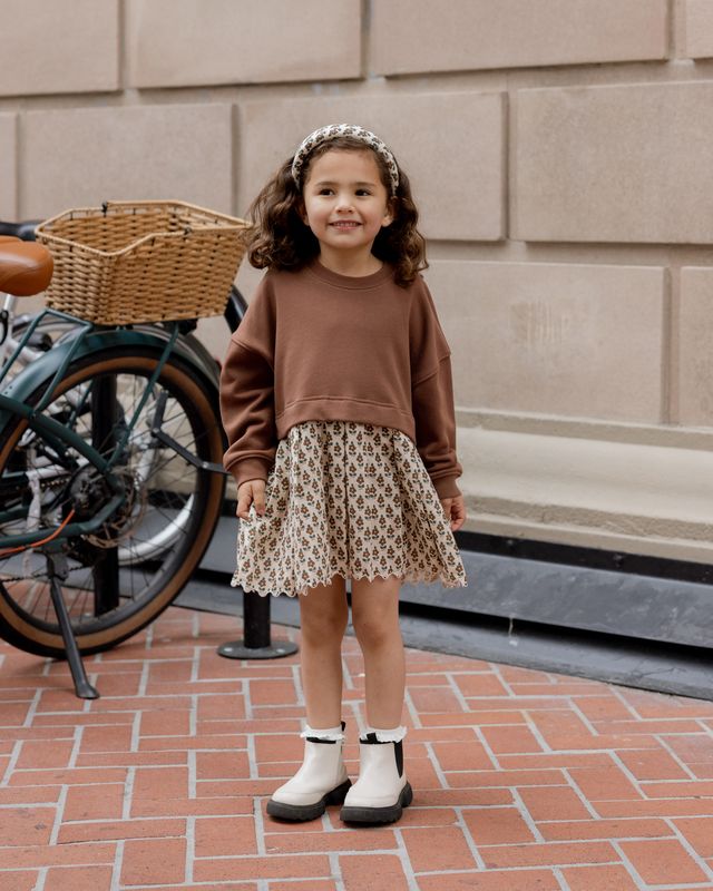 Young girl in a brown sweater and patterned skirt standing on a brick sidewalk with a bicycle in the background.