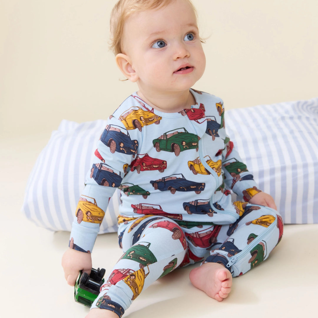 A baby sitting on a cream-colored floor or bed, wearing a light blue long-sleeve pajama set with a colorful car print. The baby is holding a small green toy car and is posed in front of a blue and white striped pillow.