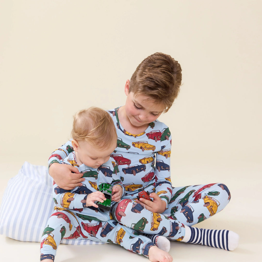Two children sitting together on a light beige studio floor, wearing matching light blue pajamas with a colorful print of toy cars. The older child has brown hair and is holding the younger child, who is looking at a small green toy.