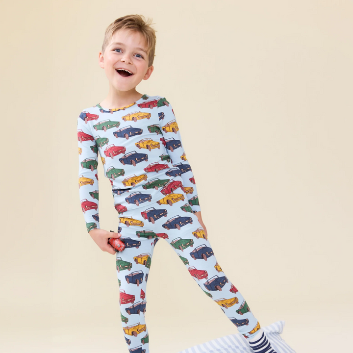 A young boy smiling and wearing a light blue long-sleeve pajama set with an all-over colorful car print, standing against a solid beige background.