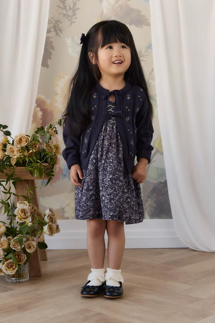 A young girl with long black hair stands indoors wearing a dark navy cardigan over a matching navy and white floral dress, paired with white socks and dark shoes. The background features white draped curtains, a floral wallpaper, and a wooden floor with a vase of roses on the left.