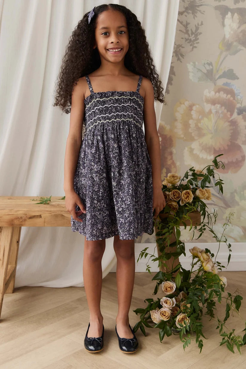 A young girl with long curly hair stands indoors wearing a sleeveless smocked dress with a dark navy and gray floral pattern, paired with dark navy flats. She is posed in front of a floral wallpaper background, a wooden bench, and a floral arrangement.