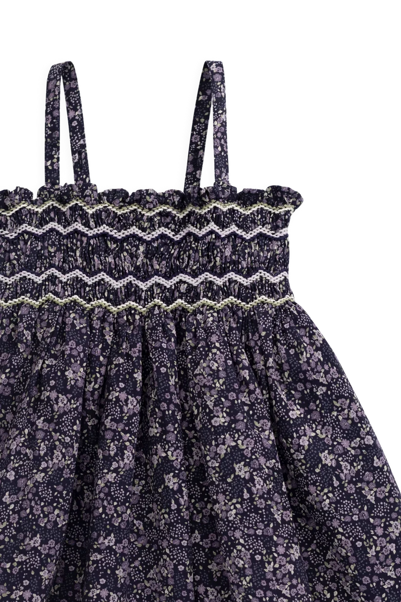 Close-up of a children's dress with thin shoulder straps, a smocked elastic waistband with embroidered zigzag trim, and a gathered skirt in a dark purple floral print.