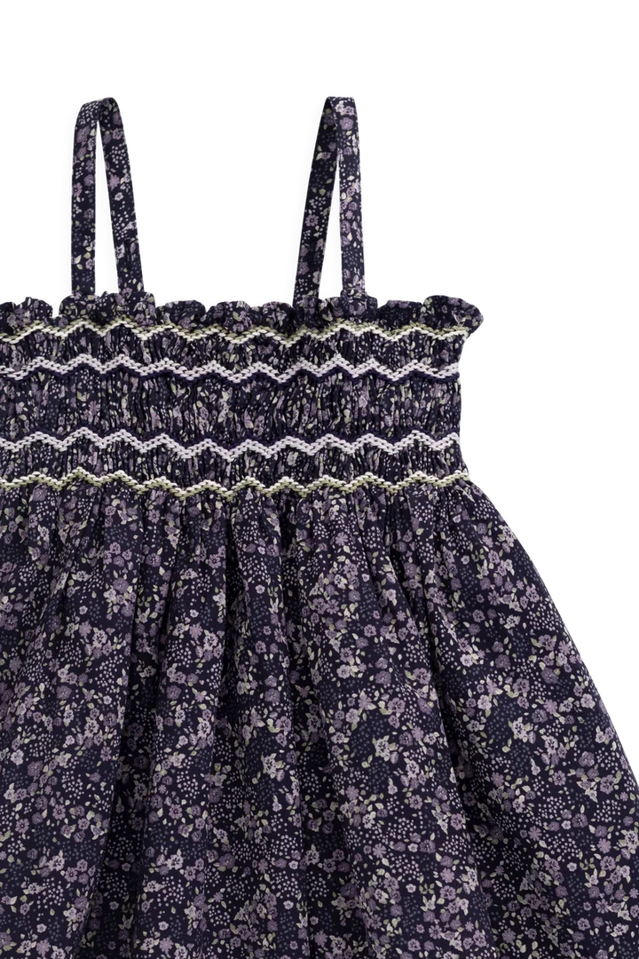 Close-up of a children's dress with thin shoulder straps, a smocked elastic waistband with embroidered zigzag trim, and a gathered skirt in a dark purple floral print.