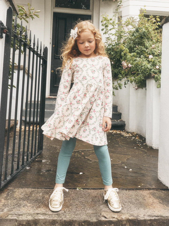 A young girl with long wavy blonde hair stands on stone steps in front of a white house, wearing a long-sleeve cream dress with a pink and green floral print, teal leggings, and white sneakers.