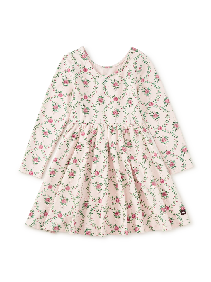 A long-sleeve baby dress with a pink and green floral print on a light cream background, featuring a gathered waist and a small brand tag near the hem.