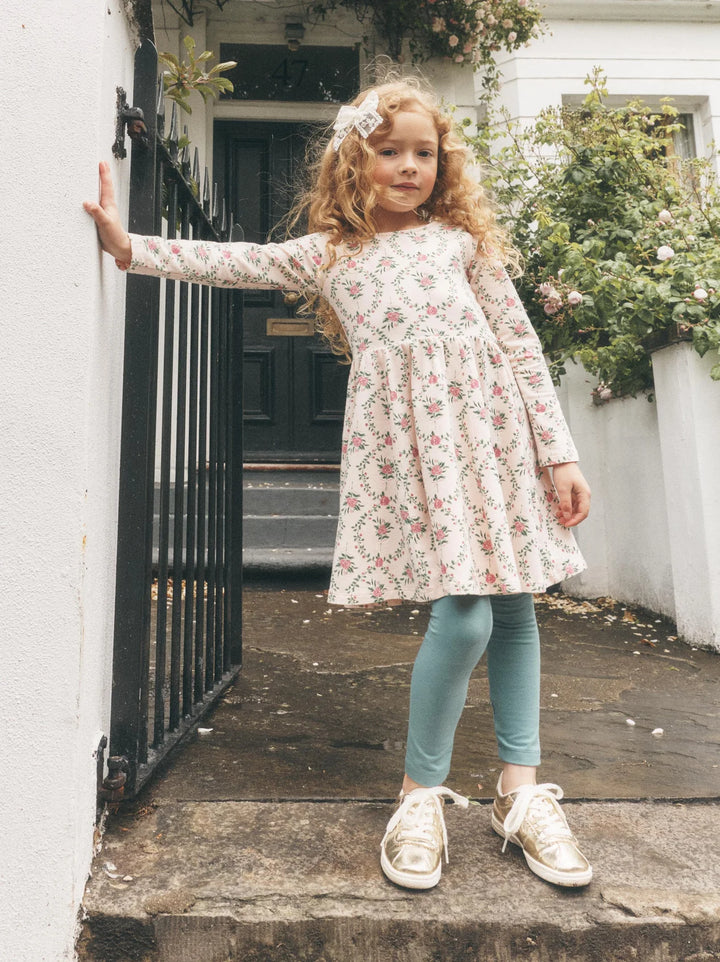 A young girl with curly blonde hair stands on stone steps in front of a white building, wearing a long-sleeve cream dress with a pink and green floral print, light blue leggings, and white sneakers.