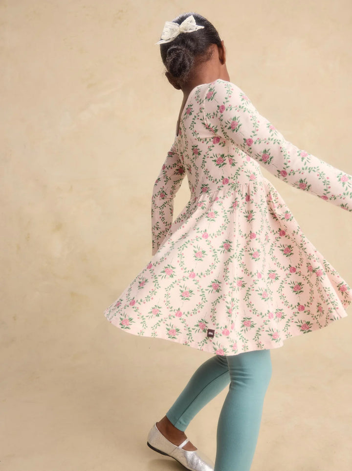 A girl wearing a cream-colored long-sleeve tunic dress with a pink and green floral pattern, paired with teal leggings and white Mary Jane shoes, captured from behind as she twirls against a textured beige background.