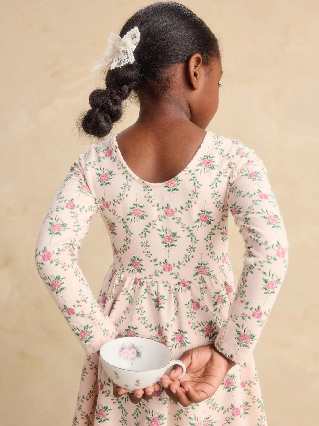 A young girl with her hair in a braid wearing a long-sleeve cream dress with a pink and green floral print, holding a small white teacup from behind.