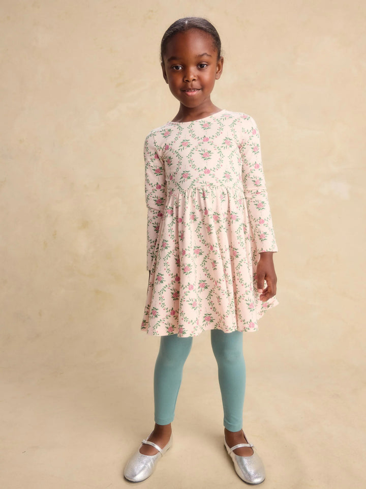 A young girl stands against a textured beige studio backdrop wearing a long-sleeve cream dress with a pink and green floral print, mint green leggings, and silver ballet flats.