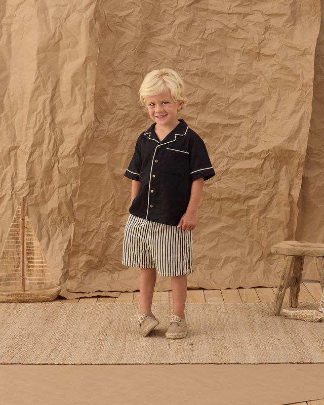 little boy wearing a black button down with white piping and a pair of black and white striped shorts