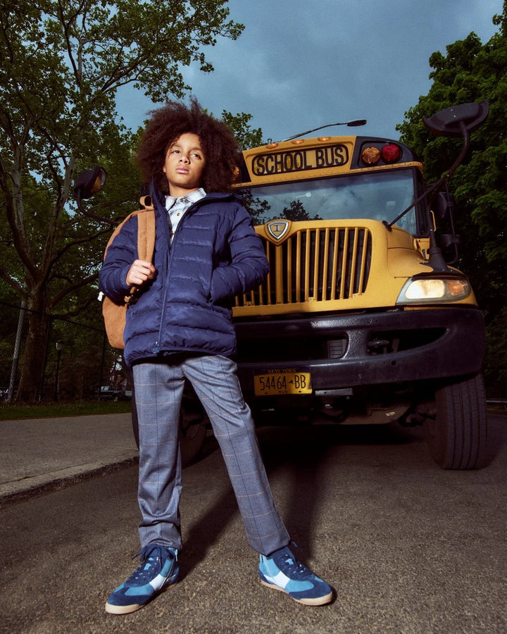 young boy standing in front of a school bus in a navy coat and gray windowpane stretch pants. 