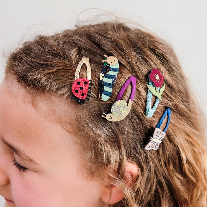 Five colorful hair clips shaped like a ladybug, striped insect, flower, and butterfly, shown attached to a child's brown hair against a light gray background.