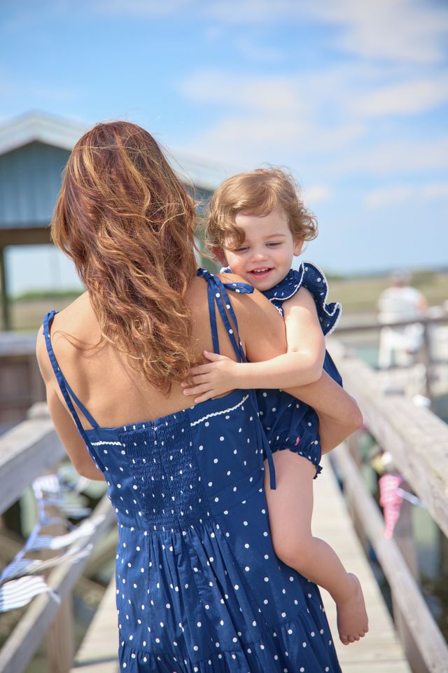 Woman in a blue dress holding a child on a wooden pier with a clear sky.