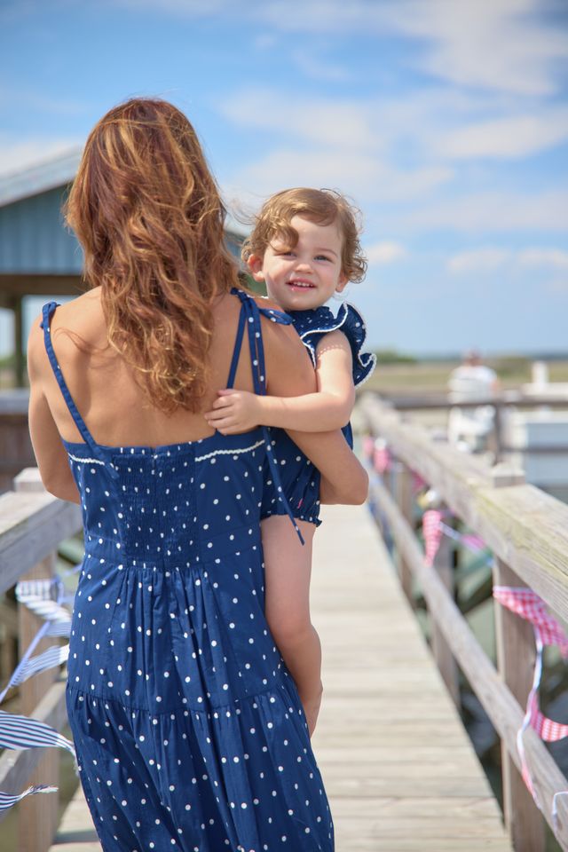 Woman in a blue polka dot dress holding a child on a wooden pier with a clear sky.