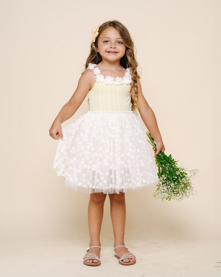 Young girl in a yellow dress with a white ruffled skirt holding greenery against a beige background