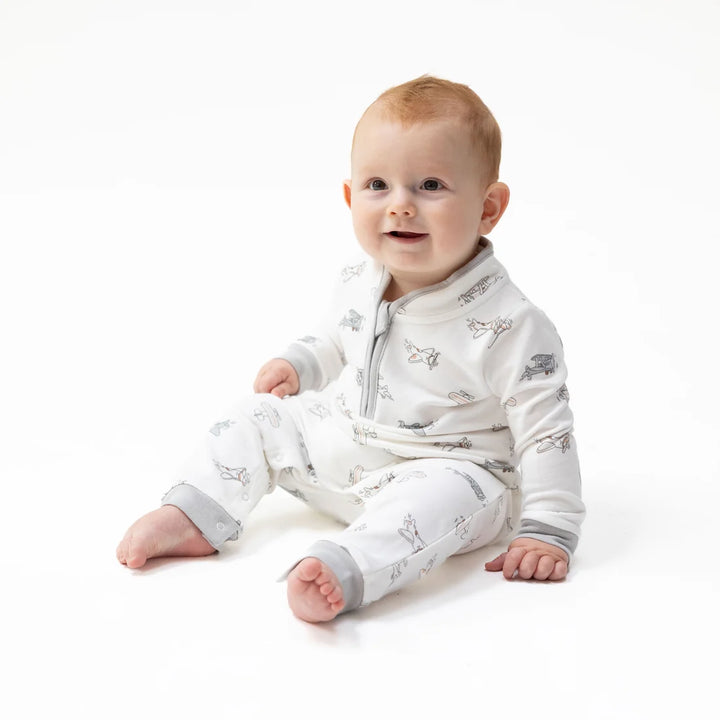 A baby sitting on a white studio background wearing a white long-sleeve footless sleeper with a gray zipper and a subtle gray vintage airplane print.